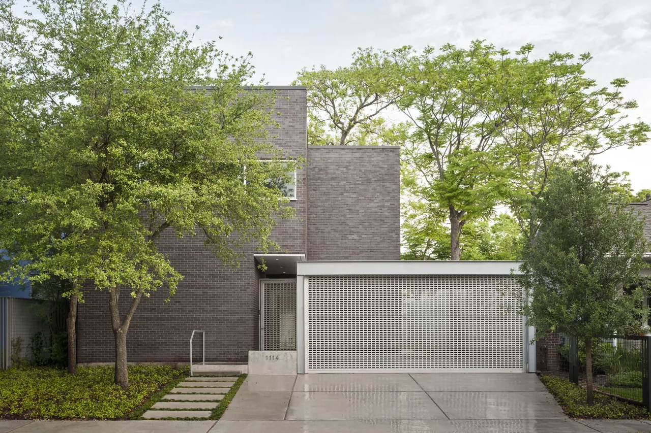 Modern brick house facade with flat roof, a perforated metal garage door, and green leafy trees surrounding the entrance.
