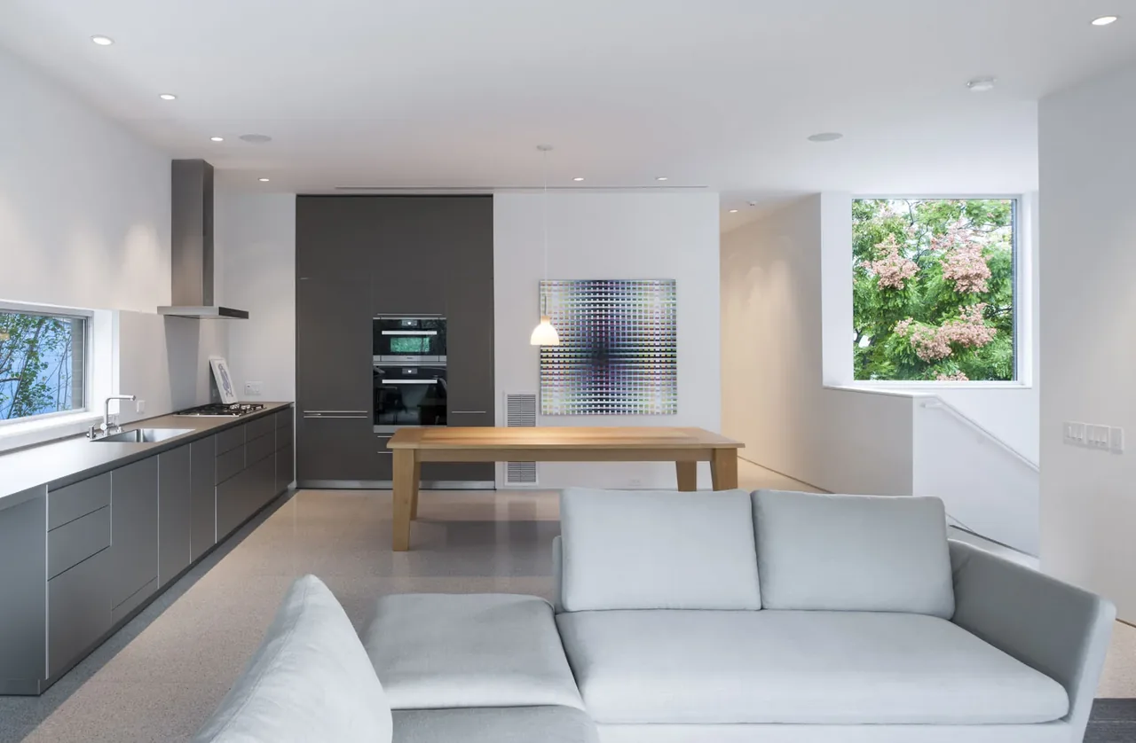 Modern kitchen and dining area in Crowley House featuring grey cabinets and a wooden dining table near a large window.