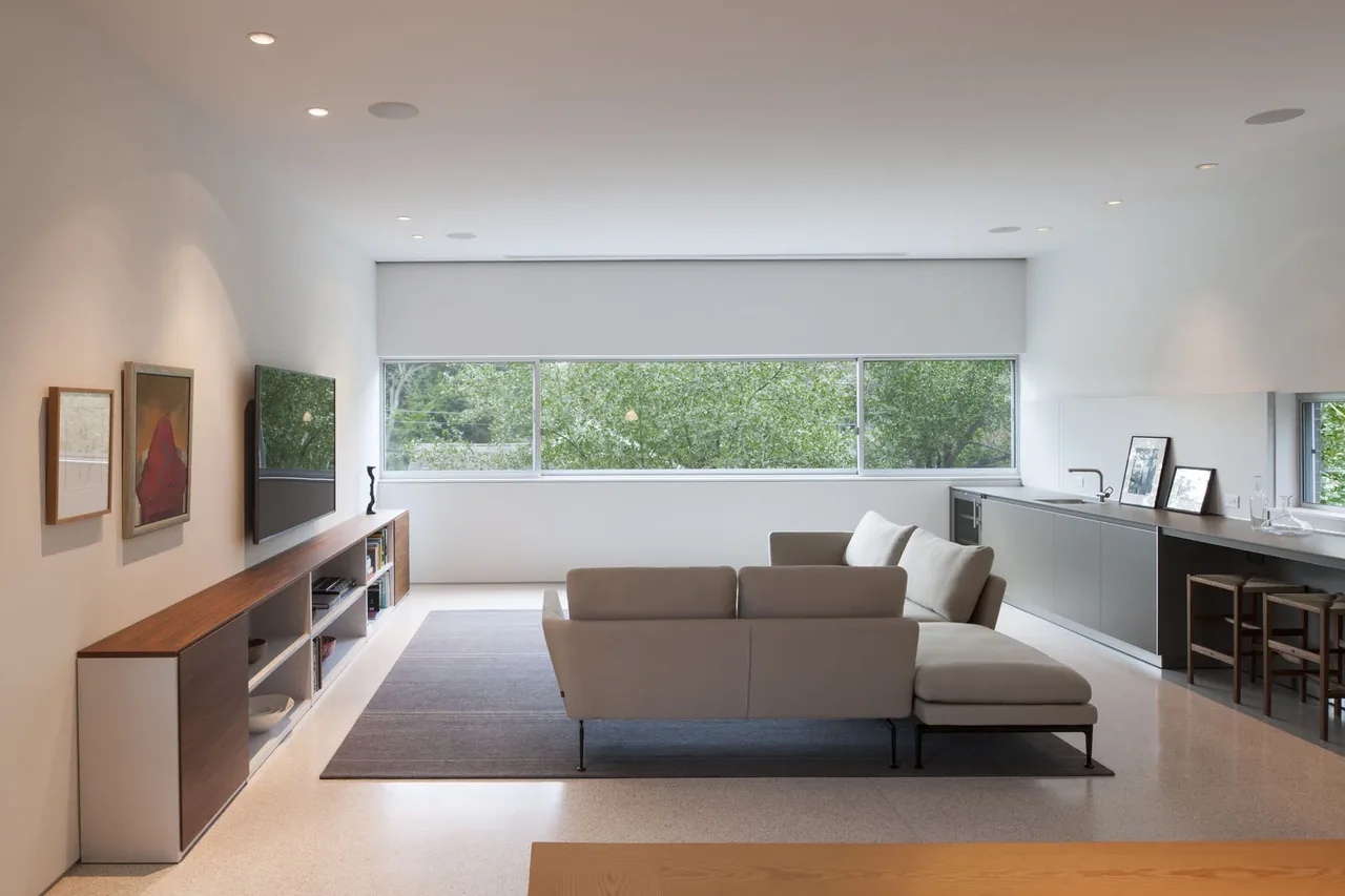 Living room in Crowley House featuring a large horizontal window, gray sectional sofa, and built-in cabinetry along the side wall.