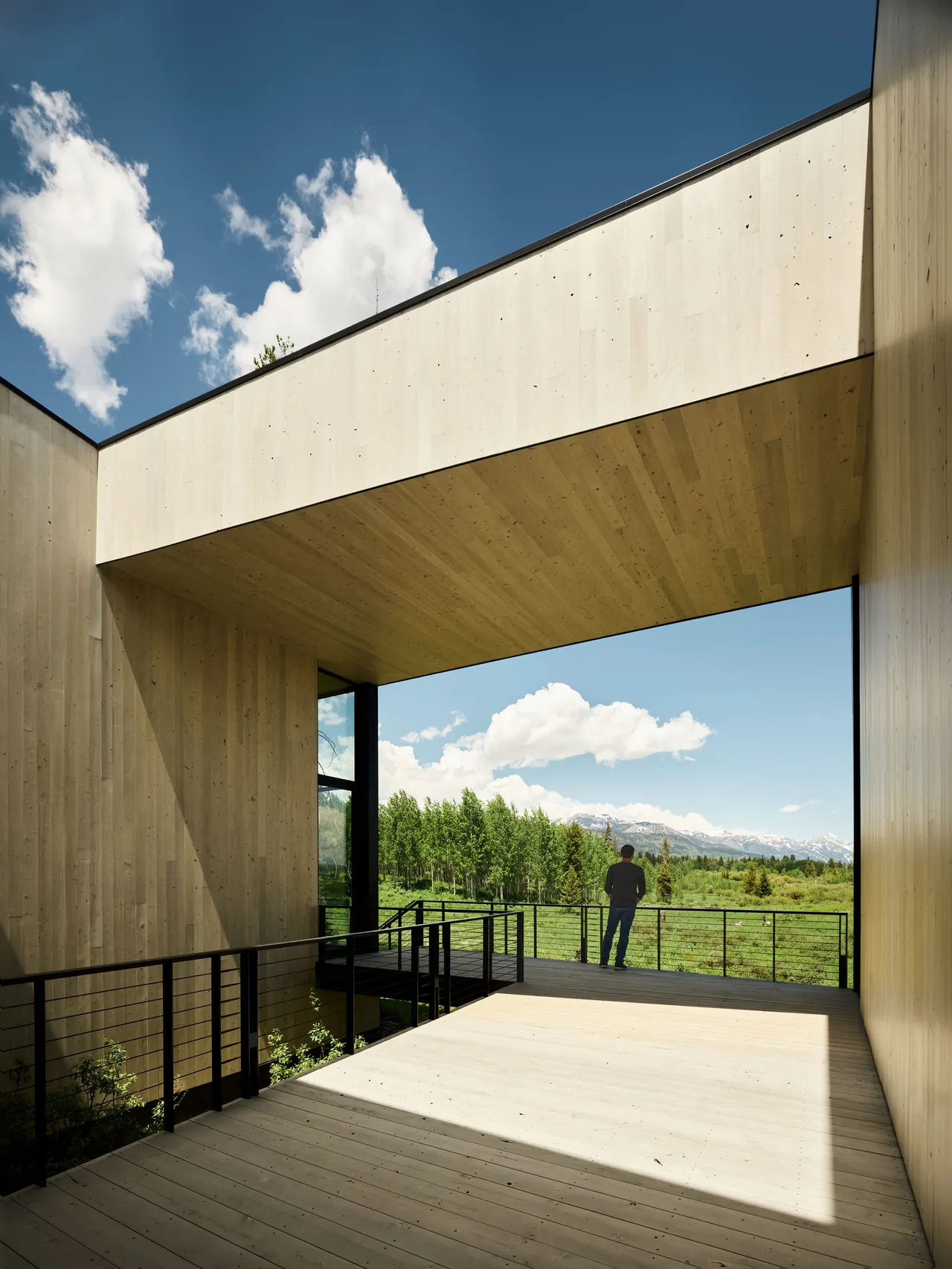 An outdoor balcony framed by light wood siding and ceiling, overlooking a green landscape with a mountain backdrop.