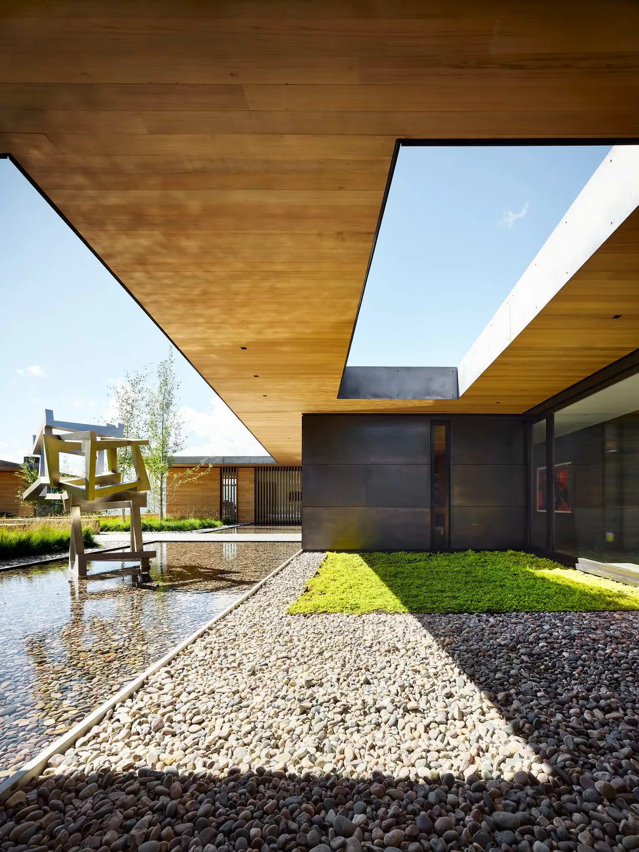 Modern courtyard with wood-clad ceiling, stone and pebble ground surface, and water feature with abstract sculpture.