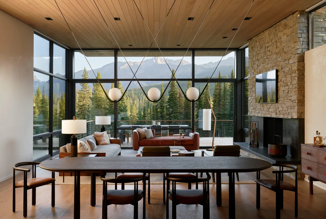 Living and dining area with large black-framed windows, stone fireplace, and wood ceiling showcasing mountain views.