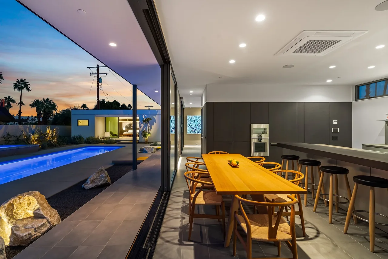 View of a modern dining room and kitchen with bar stools opening to an outdoor rectangular pool and living space at sunset.