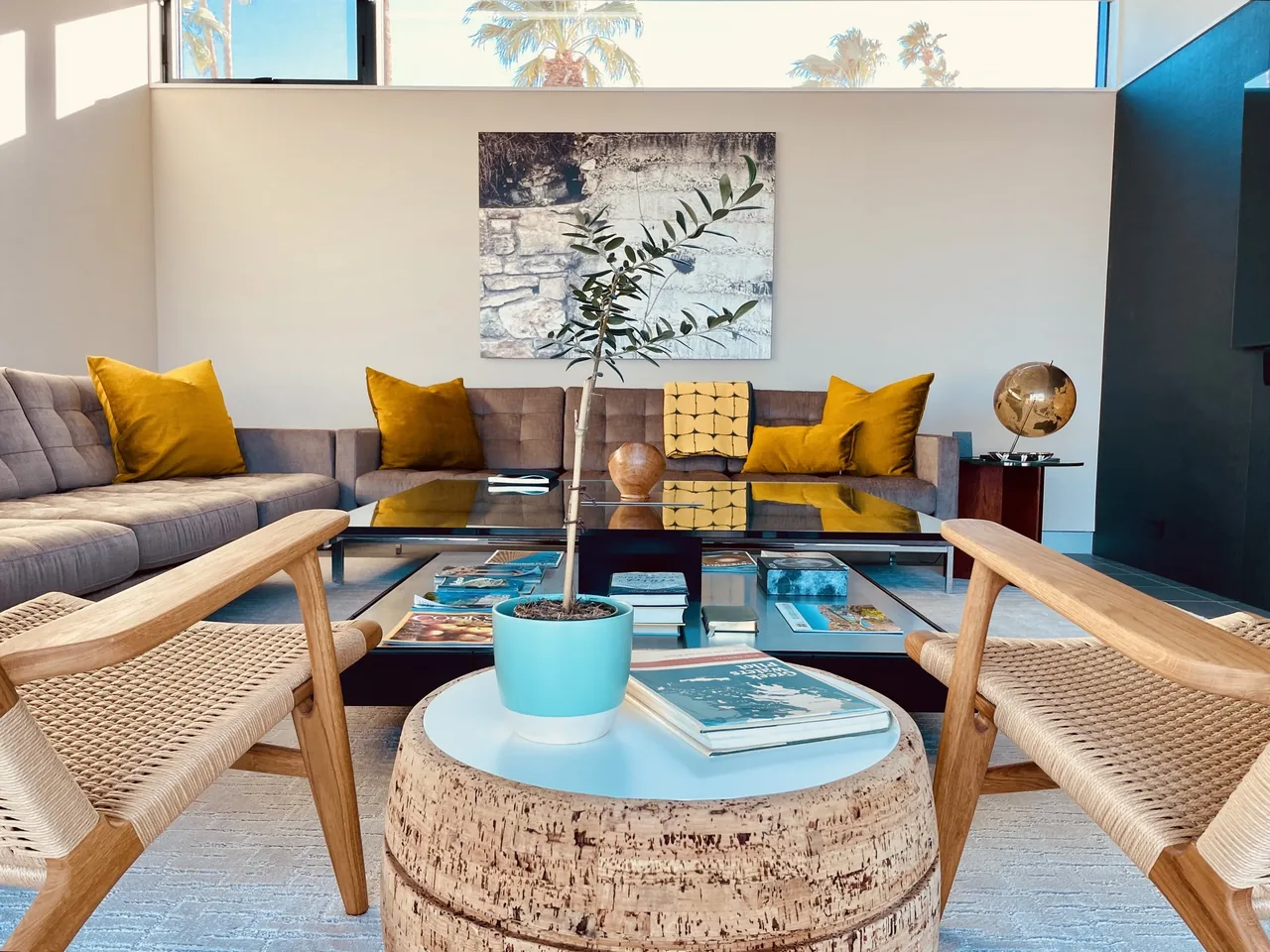 Living room featuring a gray sectional sofa with yellow pillows, wood lounge chairs, and a black glass coffee table under a horizontal window.