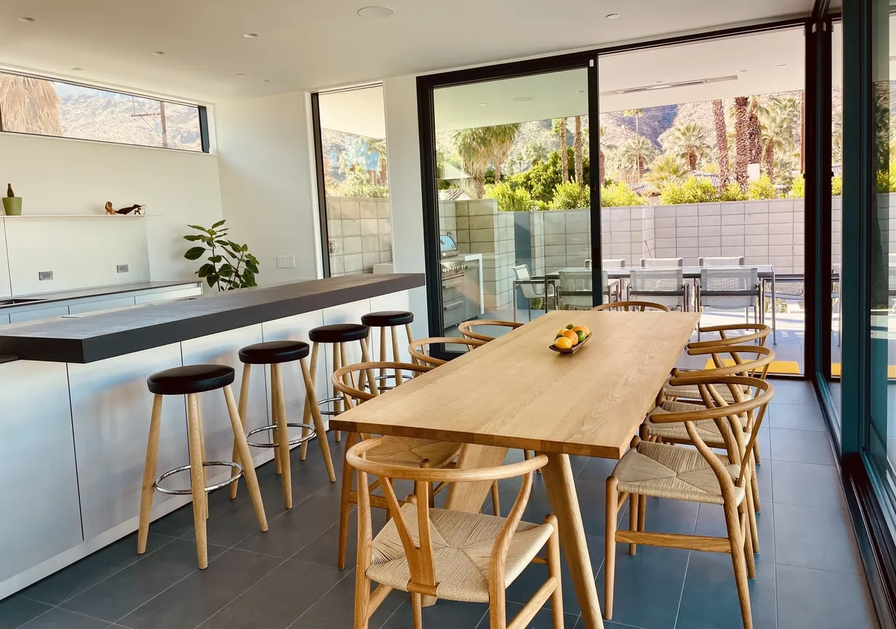 Open dining and kitchen area featuring a wooden dining table with chairs and a kitchen island with bar seating, illuminated by natural light through large sliding doors.