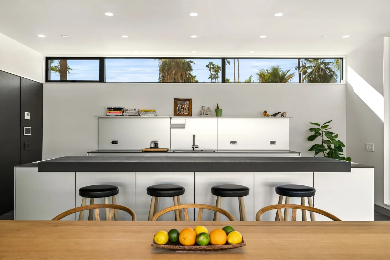 Kitchen interior with a minimalist black countertop island and a horizontal clerestory window revealing palm trees.