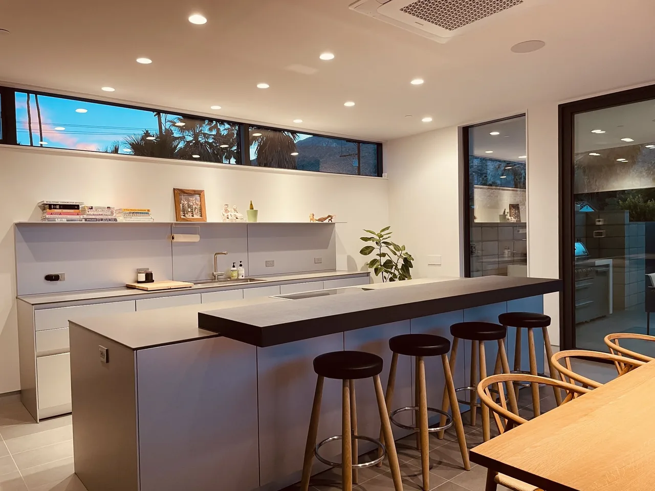 Modern kitchen with a sizable island bar with stools, overlooking a dining table and outdoor area at dusk.