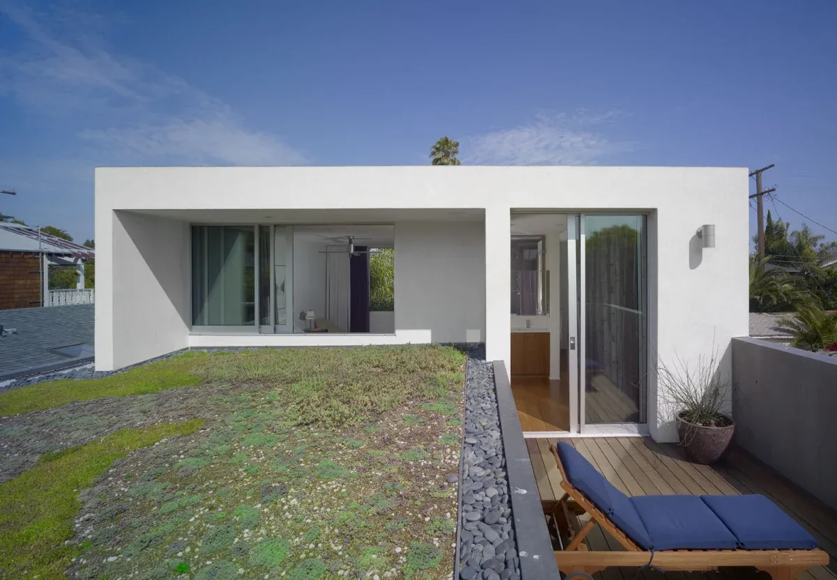 The NOWITA project features a contemporary white exterior with a green flat roof and an elevated porch with patio furniture.
