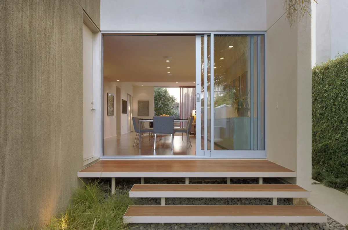 Small front porch with wooden steps and large sliding glass door showing interior dining room.