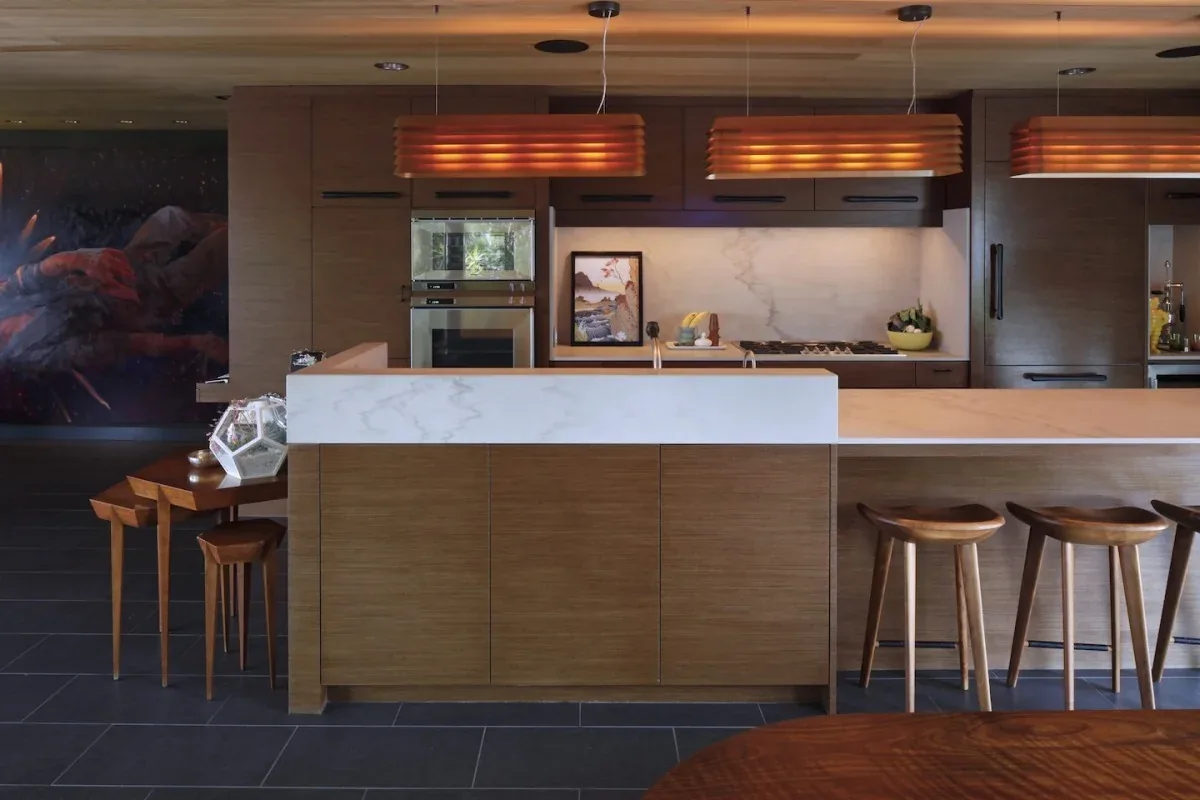 The LIVE OAK kitchen features wood cabinetry, a white marble countertop bar, and pendant lighting from Carson Architects.