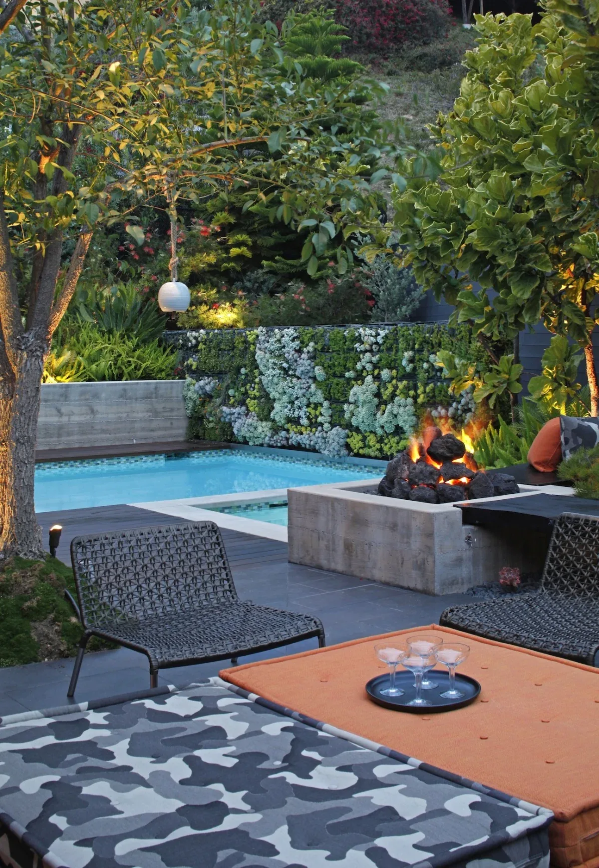 Poolside with modern outdoor furniture, fire pit, and vertical succulent garden on concrete wall at dusk.