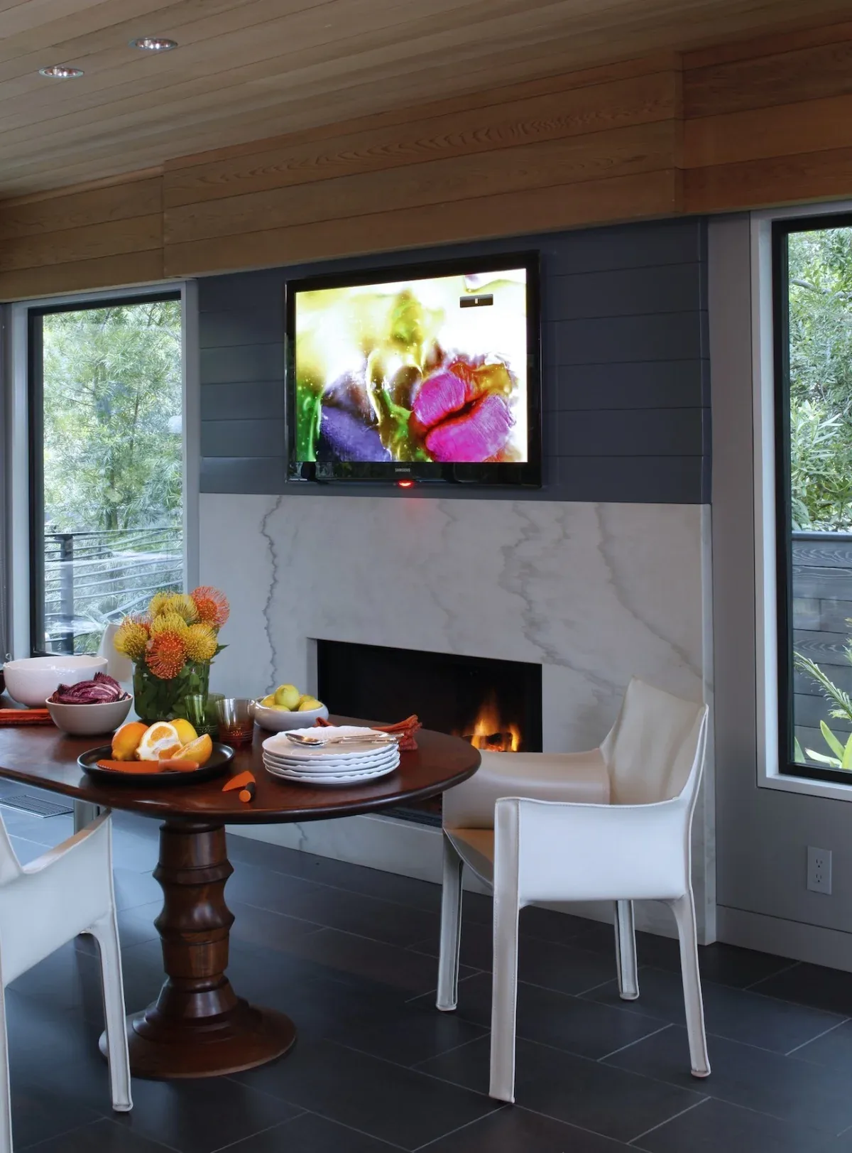Dining area features a fireplace with marble surround and TV mounted above, flanked by large windows with views of greenery outside.