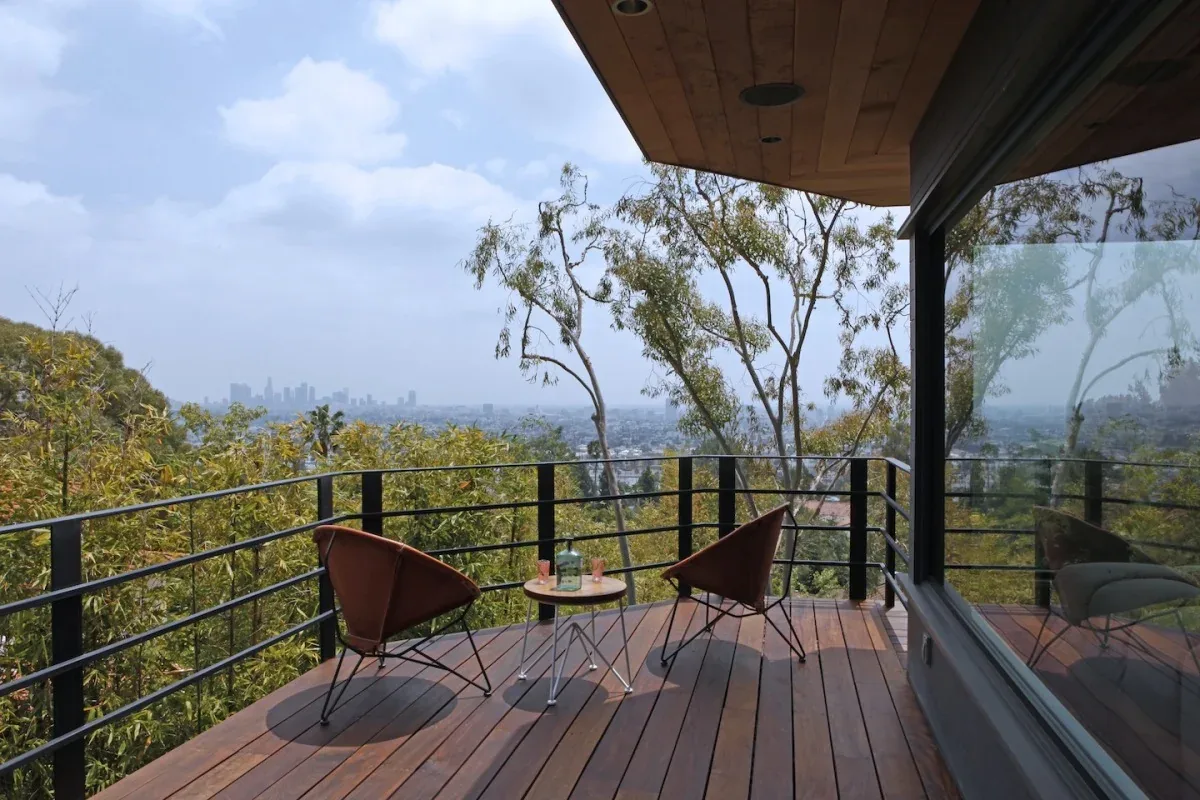 Outdoor wood deck with brown woven chairs and a small round table offers city views beyond trees.