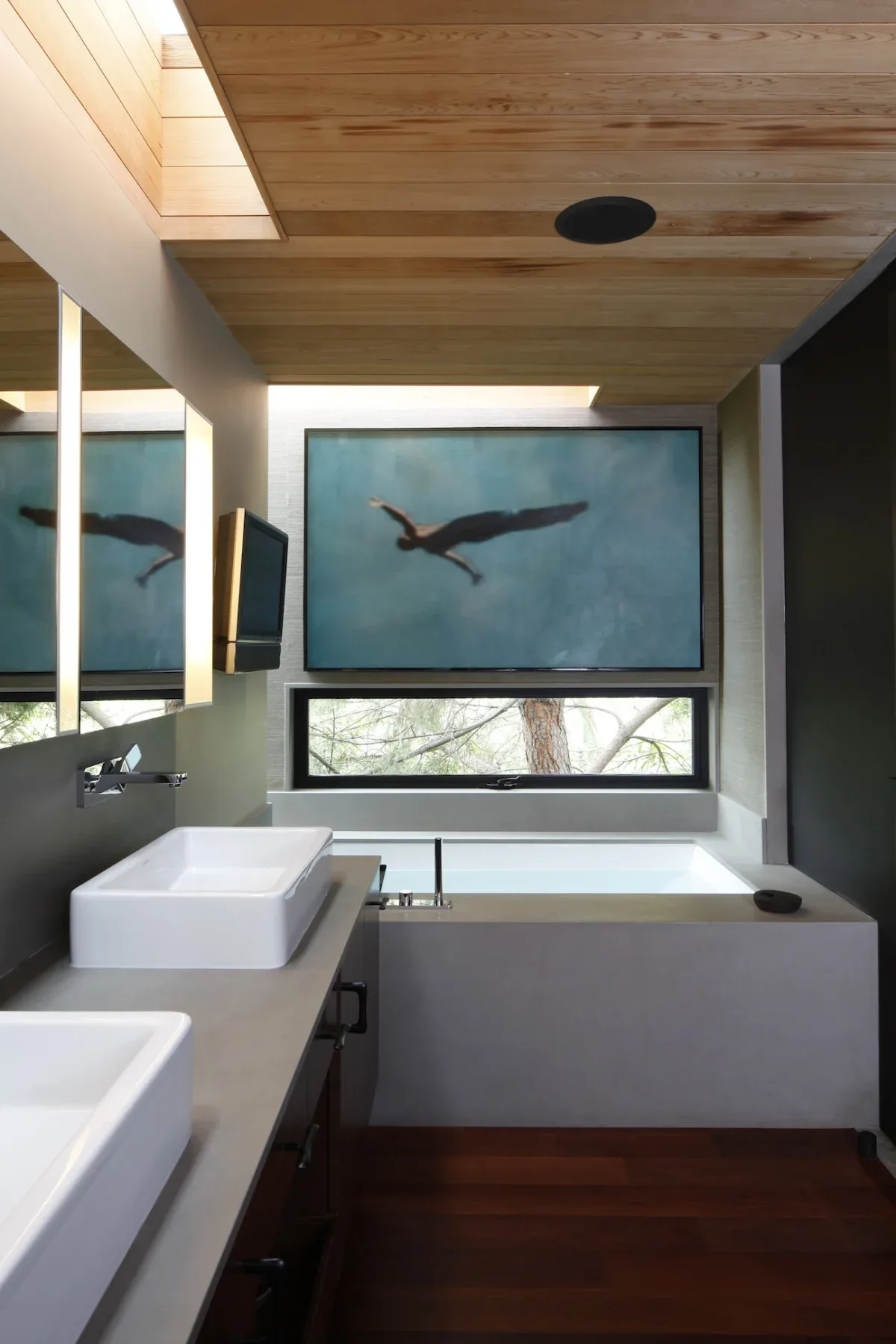 Bathroom featuring wood-paneled ceiling, rectangular white vessel sinks on a concrete countertop, and a bathtub below a horizontal window and artwork.