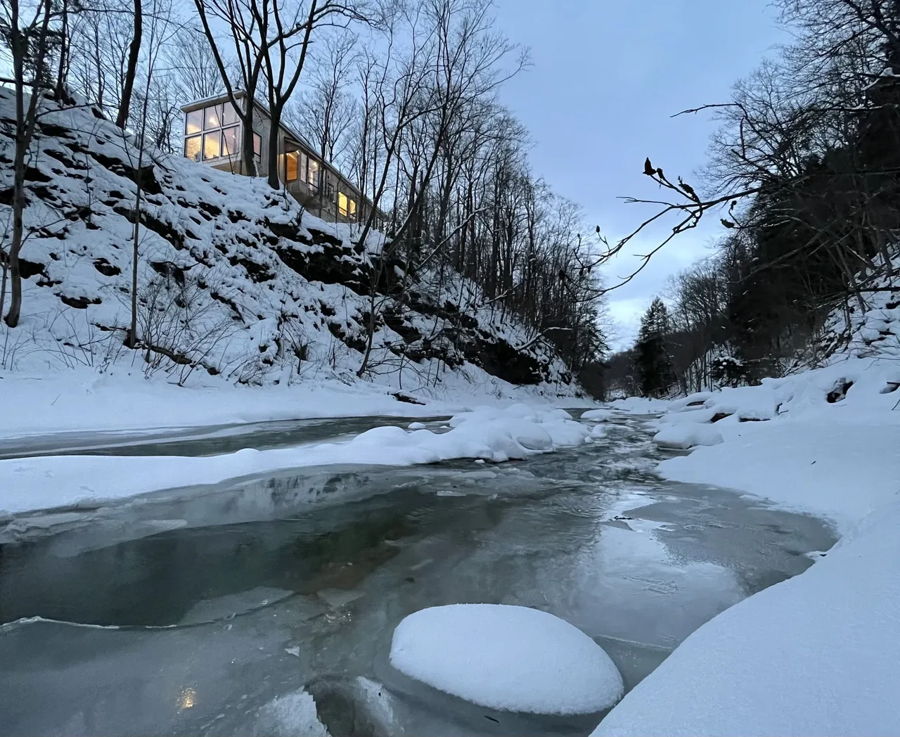 Canadaway House by Carter + Burton sits on a rocky snow-covered hill with floor-to-ceiling glazing overlooking a winter creek.