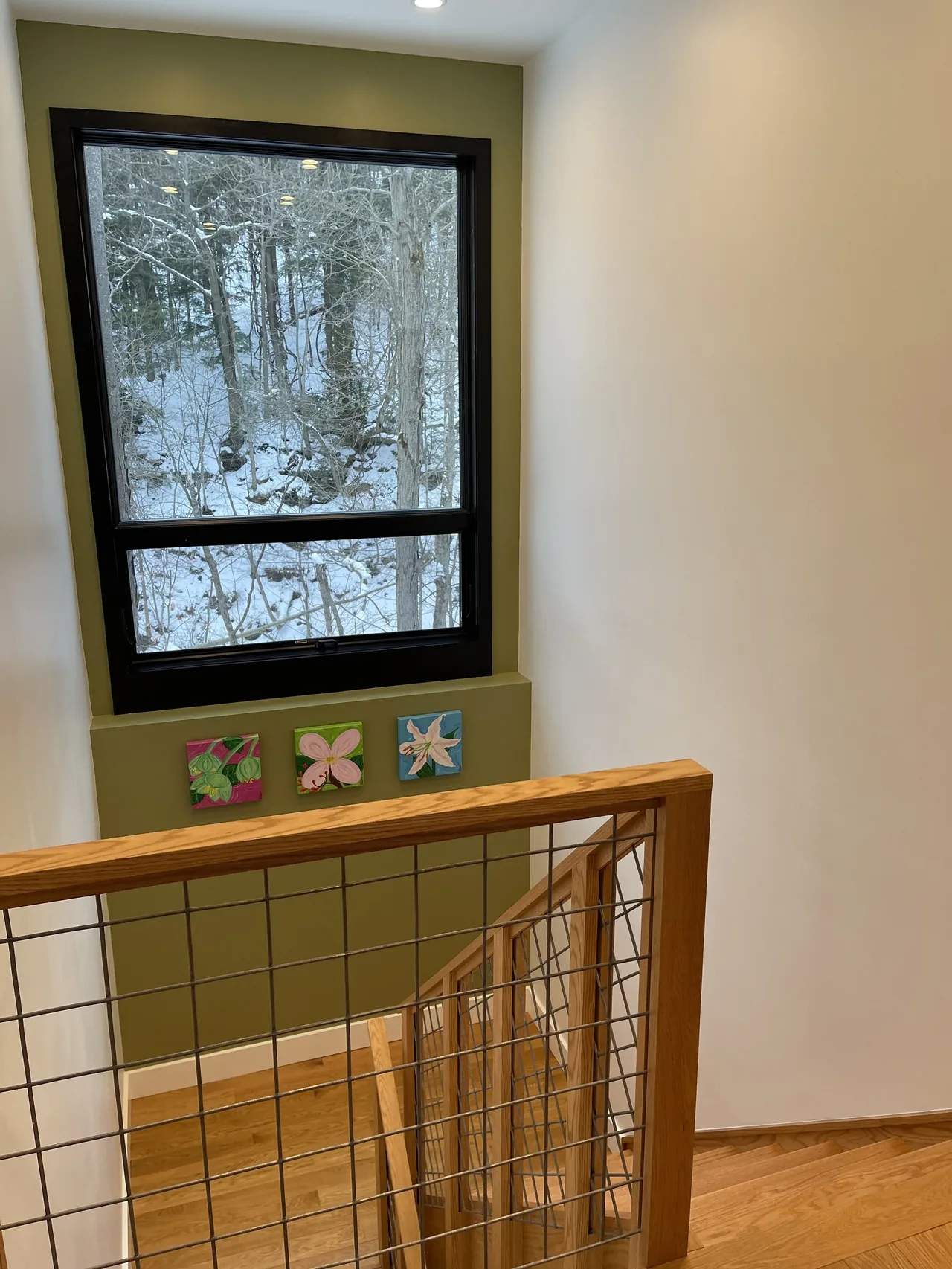 Interior stairwell with wood railing and grid wire balustrade next to a black-framed window overlooking a snowy landscape.