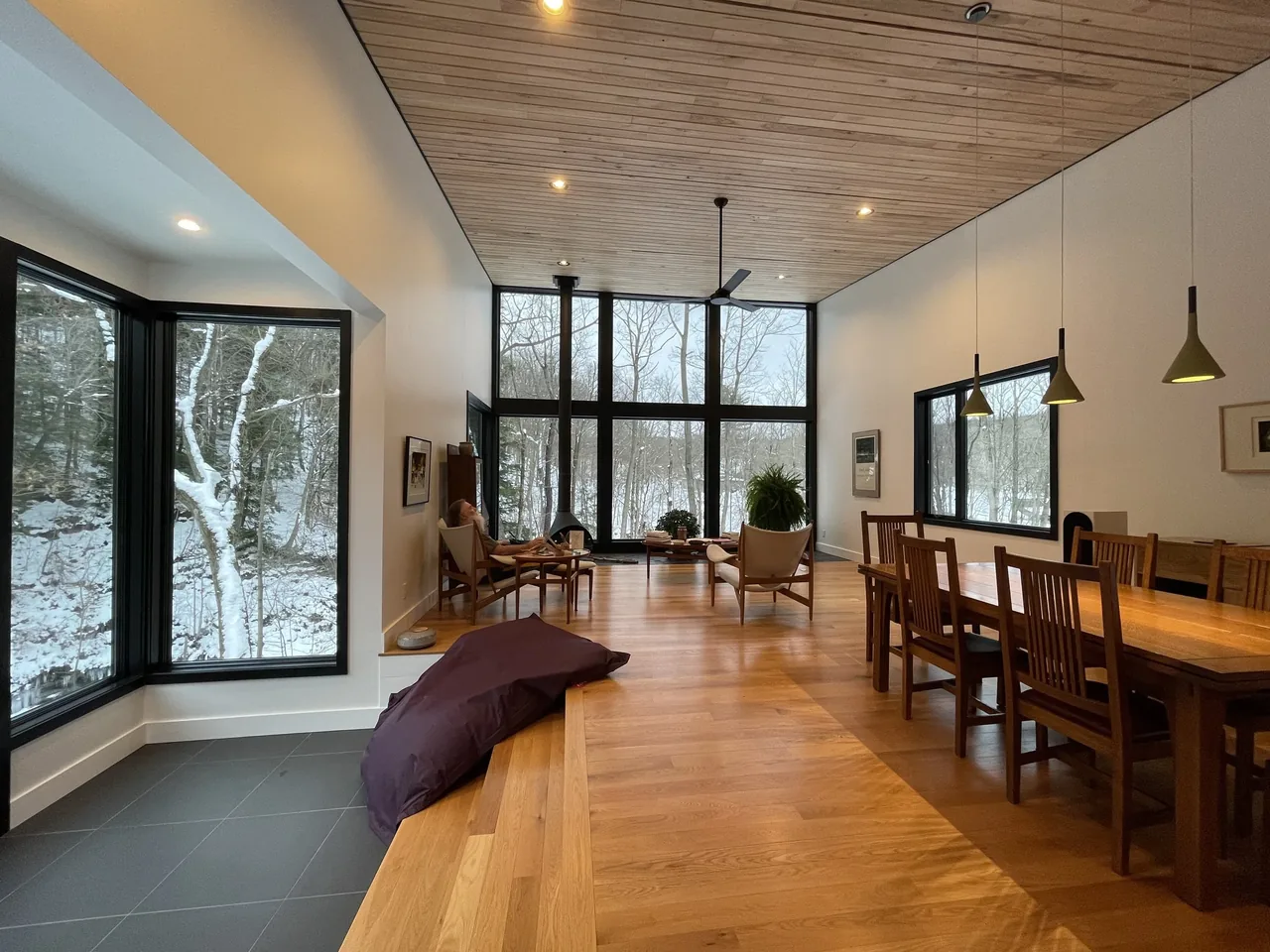 Living and dining room with wood ceiling and floors, black window frames, and large windows overlooking a snowy wooded landscape.