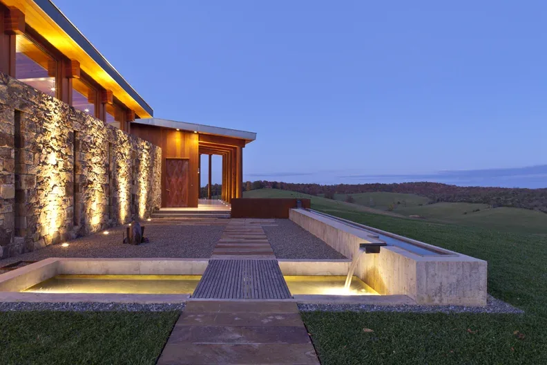 The Elk Run Ridge house features stone walls illuminated by warm lighting and integrates water elements with a scenic rural backdrop.