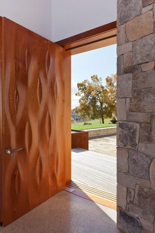 View through a textured wooden door into an outdoor patio area framed by stone walls and autumn foliage.