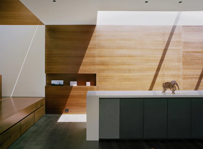 Interior detail of a wood-paneled wall with built-in shelving and minimalist cabinetry illuminated by skylight shadows.