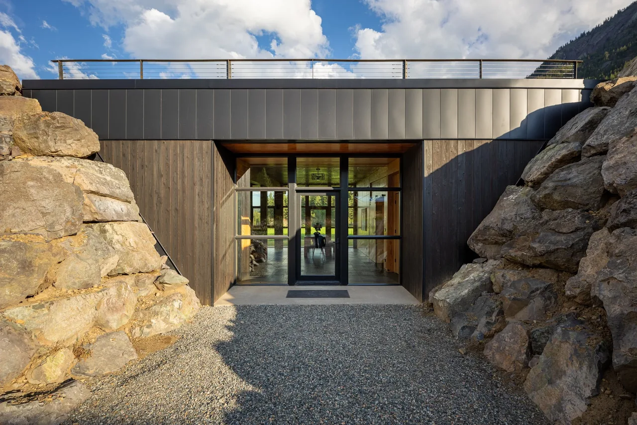 Modern berm house entrance by CAST architecture, featuring black metal and wood siding nestled between large rock walls.
