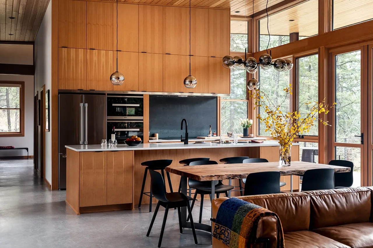 The Edelweiss House kitchen and dining space feature warm wood paneling, black accents, and expansive windows looking out to trees.