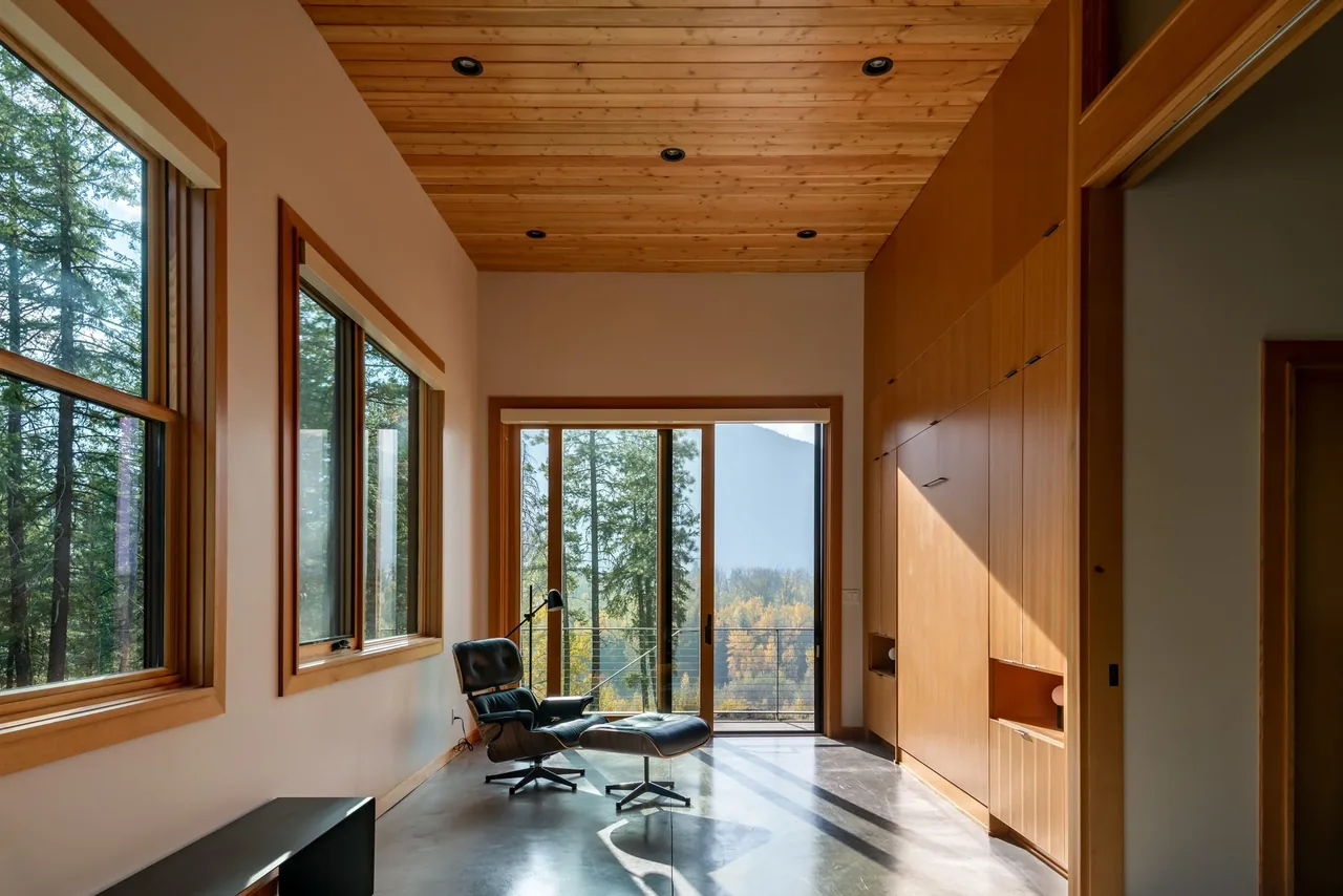 Edelweiss House seating area features wood paneling, concrete floor, and large windows overlooking forest.