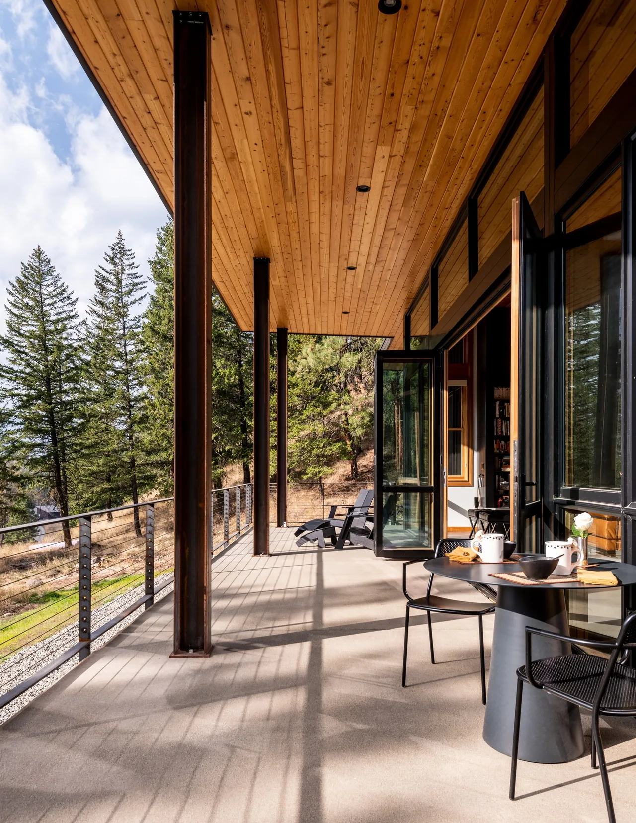 The Edelweiss House features a covered outdoor living space with wood-paneled ceiling and steel columns overlooking a natural wooded hillside.