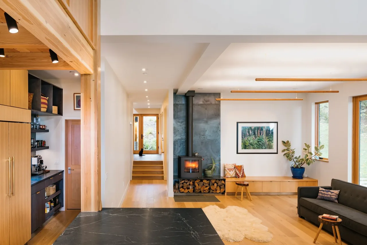 Modern living room with wood stove, built-in bench seating, and large windows showing greenery outside.