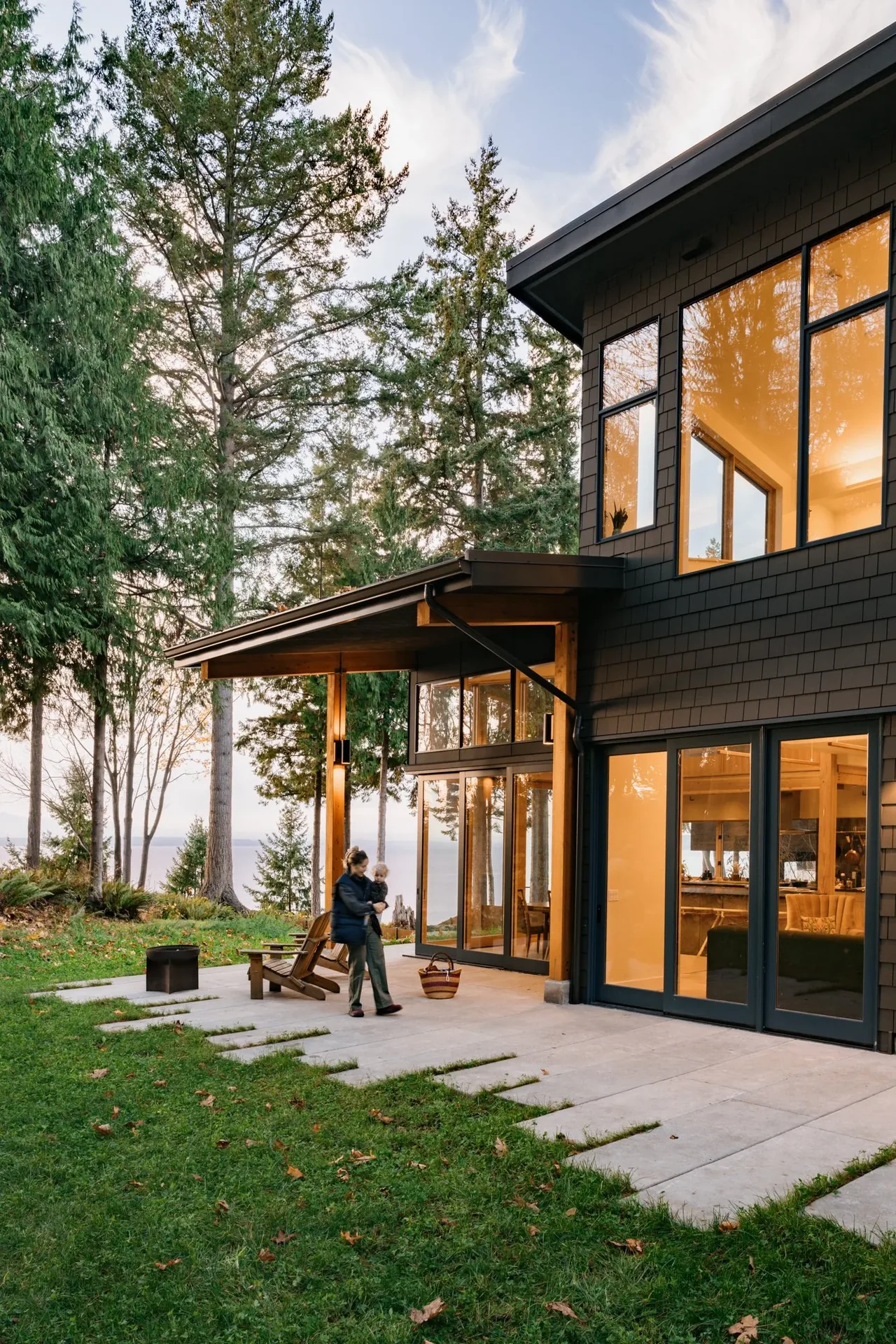 Modern two-story home with large black window frames and a covered patio overlooking a forest setting.
