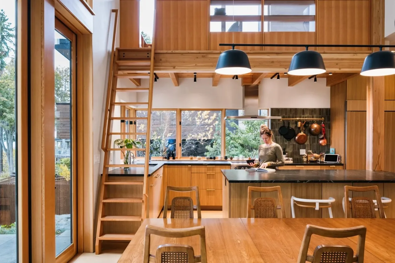EDMONDS OVERLOOK kitchen features wood cabinetry, black countertops, a wooden ladder to a loft, and large windows with forest views.