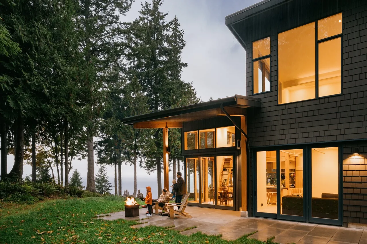EDMONDS OVERLOOK features a modern exterior with tall windows and an outdoor firepit seating area overlooking a forest setting.