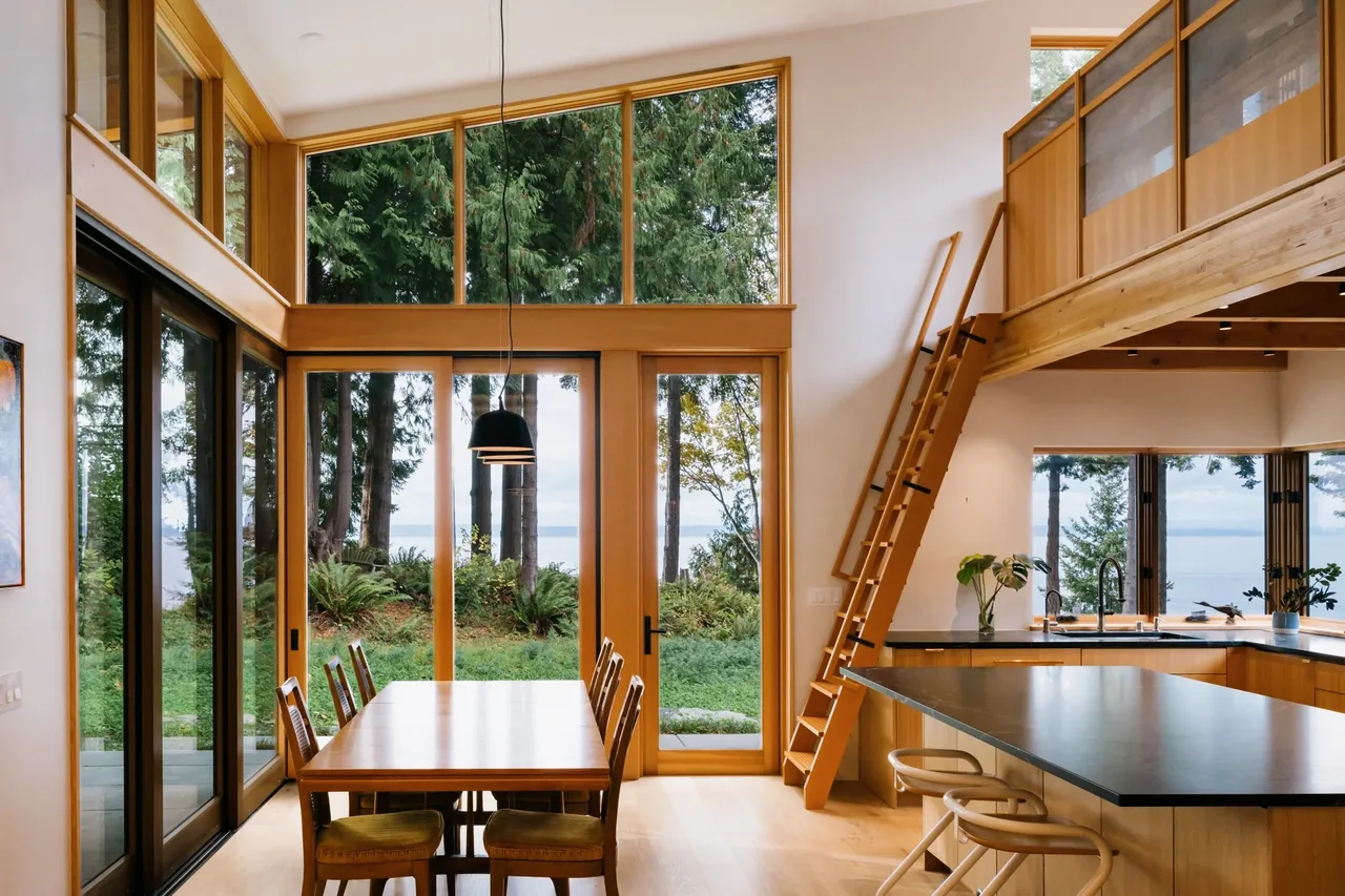 EDMONDS OVERLOOK features a dining and kitchen space with wood accents, large glazing, and a wooden ladder to the loft, framed by forest and ocean views.