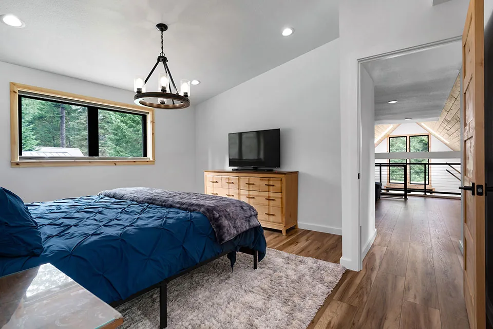Bedroom features blue bedding and natural wood furniture, connecting to a bright hallway with vaulted ceiling details.