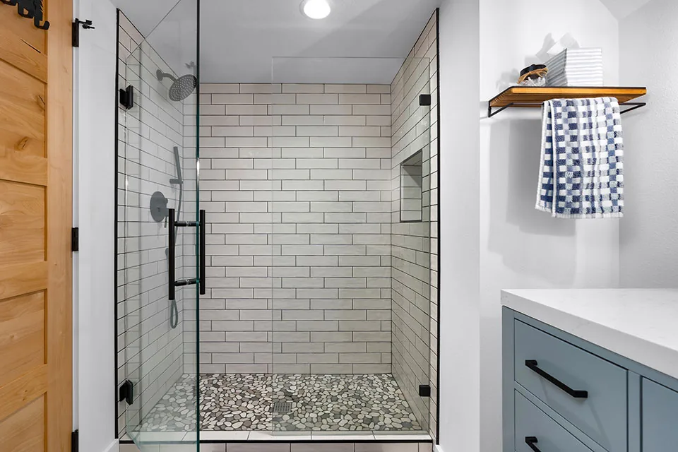 Bathroom with walk-in shower featuring subway tile and pebble stone floor, blue vanity, and wooden towel shelf.