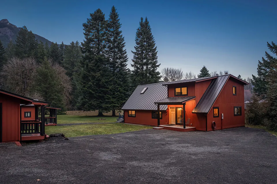 Two-story red house features a steep metal roof and a small porch, situated near tall evergreen trees in a spacious lot.