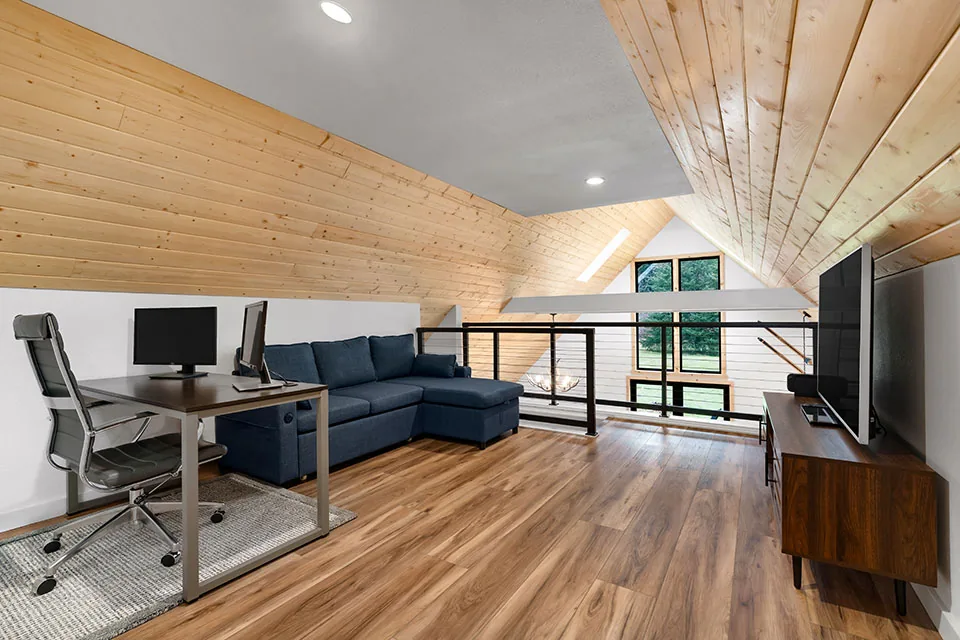 Loft space with wood-paneled sloped ceiling, hardwood floor, sectional sofa, desk setup, and large windows overlooking greenery.