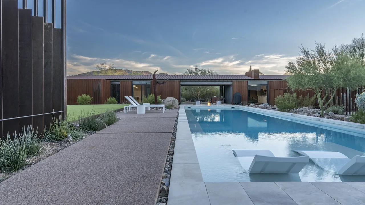 A modern single-story house with a rectangular pool and side loungers set in desert landscaping.