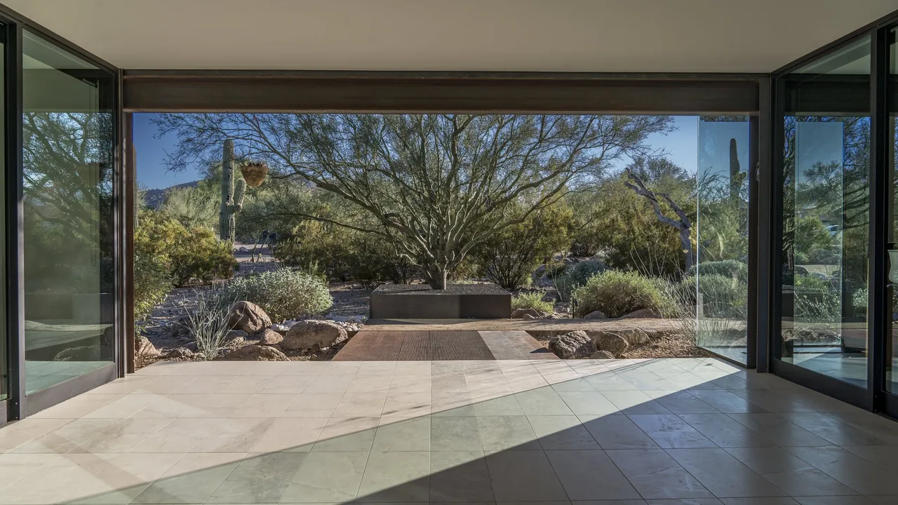 Interior view looking out through open sliding glass doors to a desert landscape with native plants and rocks.