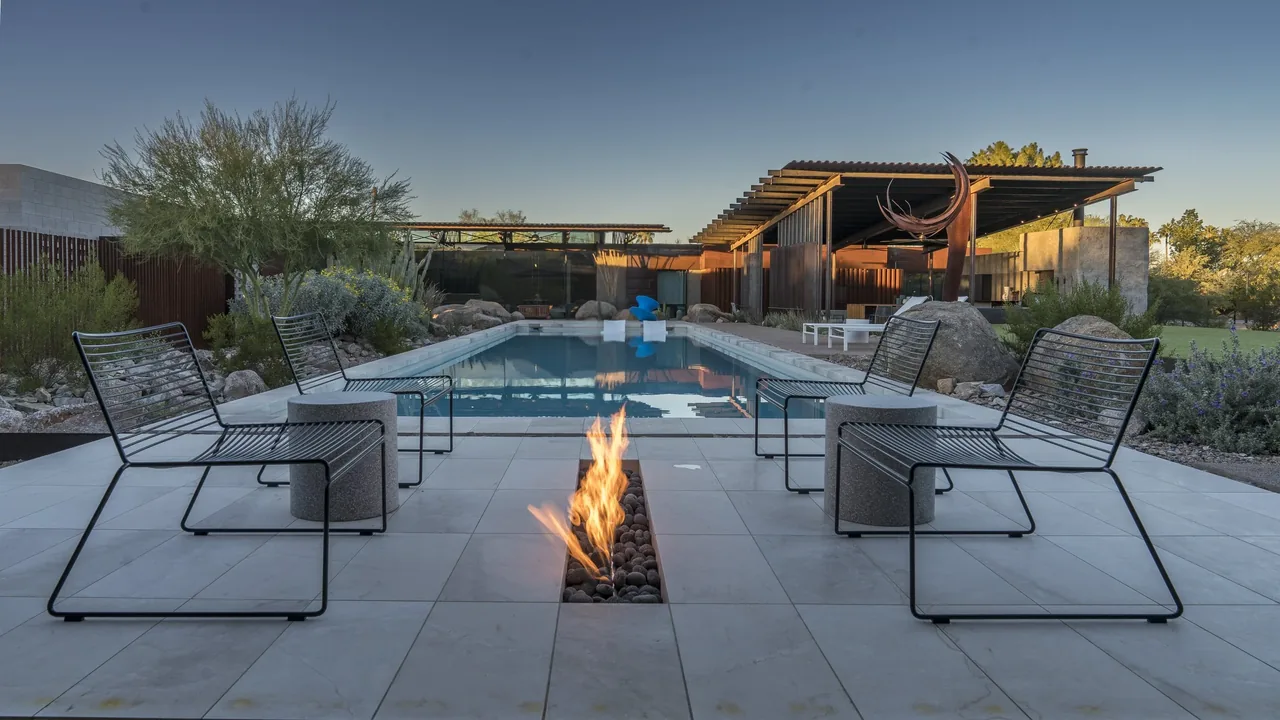 A rectangular pool features a built-in linear fire pit with metal chairs arranged on a tiled patio in a modern desert landscape.
