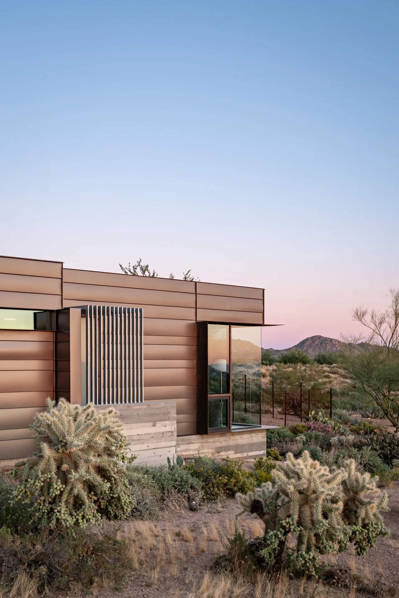 Modern home exterior with metal siding and large glass window situated in a desert landscape with mountainous backdrop.