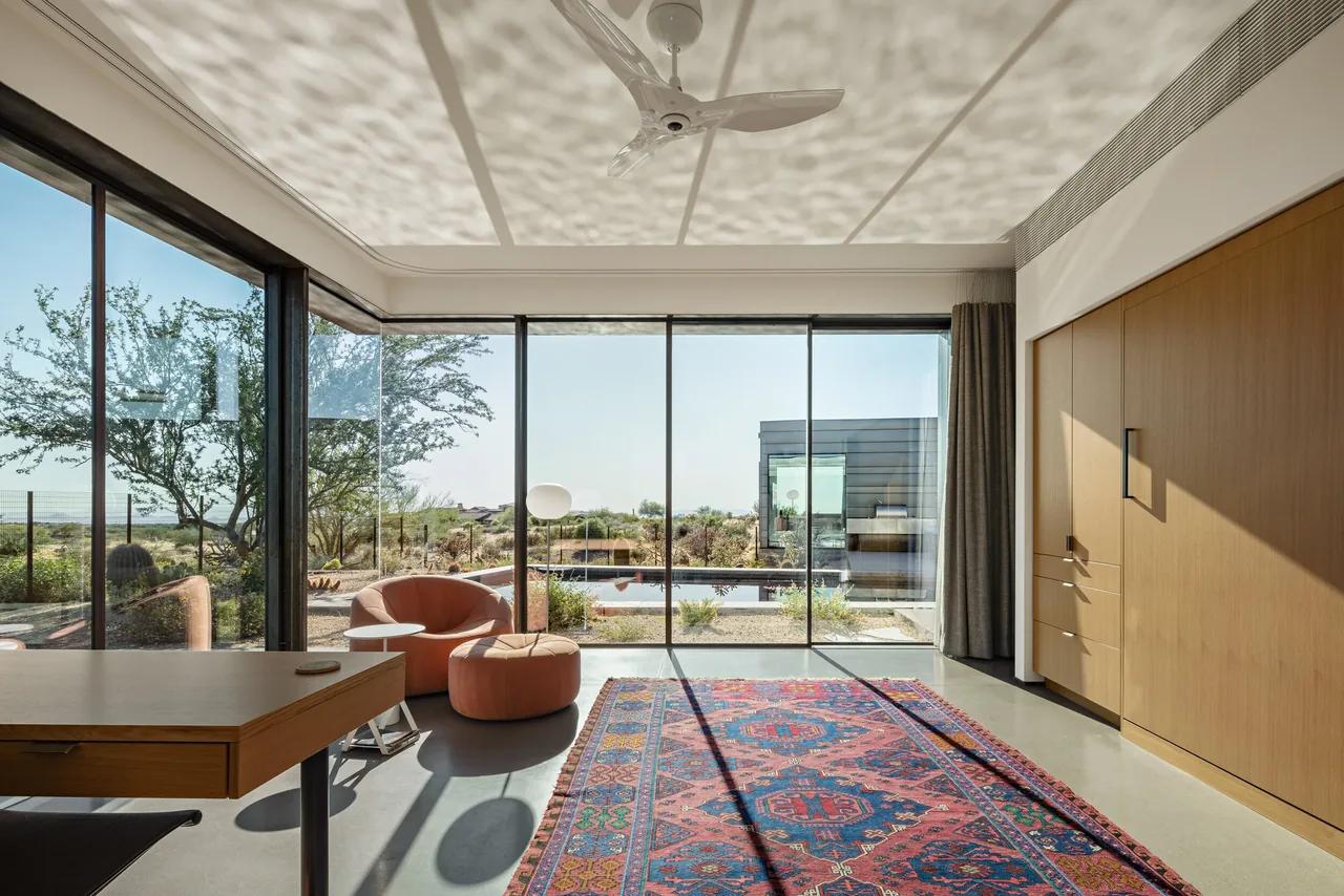 Room with floor-to-ceiling glazing overlooking desert and pool, featuring natural wood storage and a colorful rug.