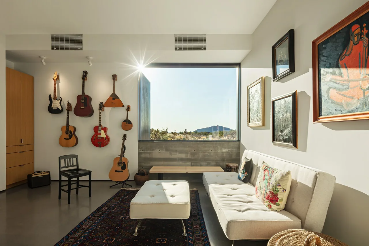 Living room with multiple guitars on wall, a beige tufted sofa with floral pillows, an ottoman on a patterned rug, and large window framing desert view.