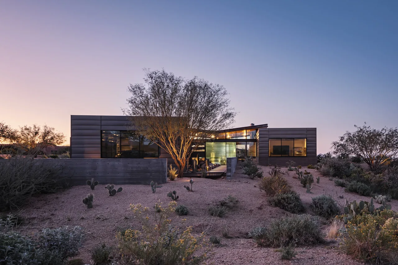 Modern metal-paneled house in a desert setting with large windows and integrated outdoor lighting during twilight.