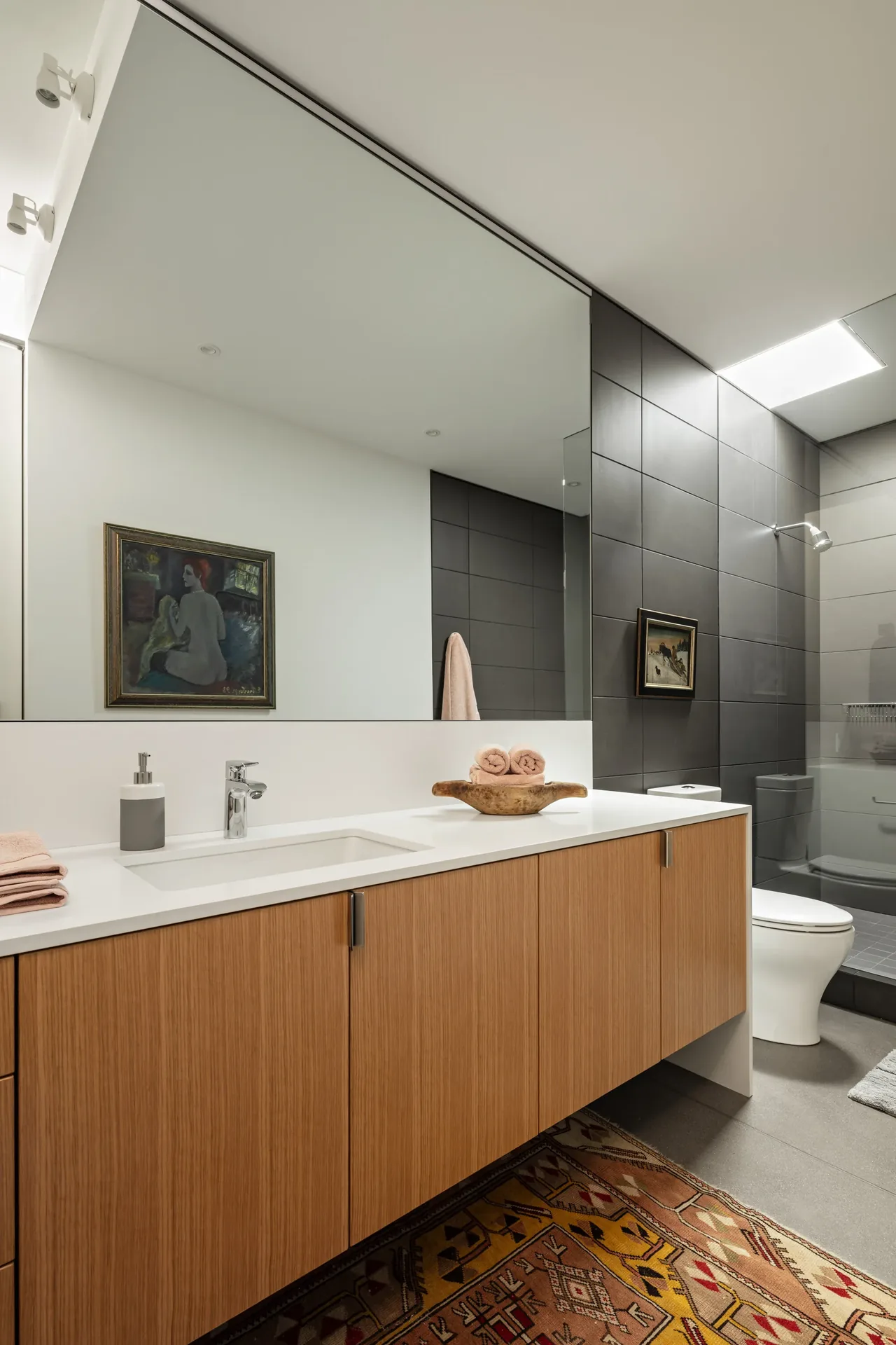 Bathroom features a floating wood vanity, large mirror, black tiled shower wall, and a colorful patterned rug.
