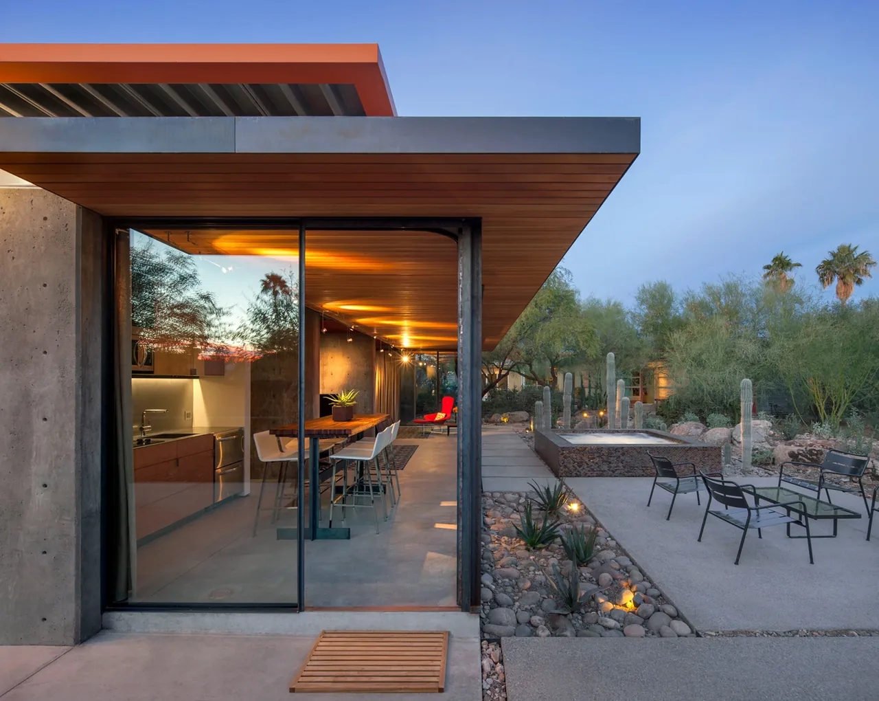 Modern barn guest house features floor-to-ceiling glazing opening to a desert landscaped patio with outdoor seating and reflecting pool.