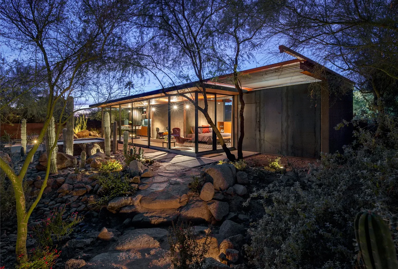 The barn guest house features floor-to-ceiling glazing and a flat roof, nestled in a desert landscape with cacti and rocks.
