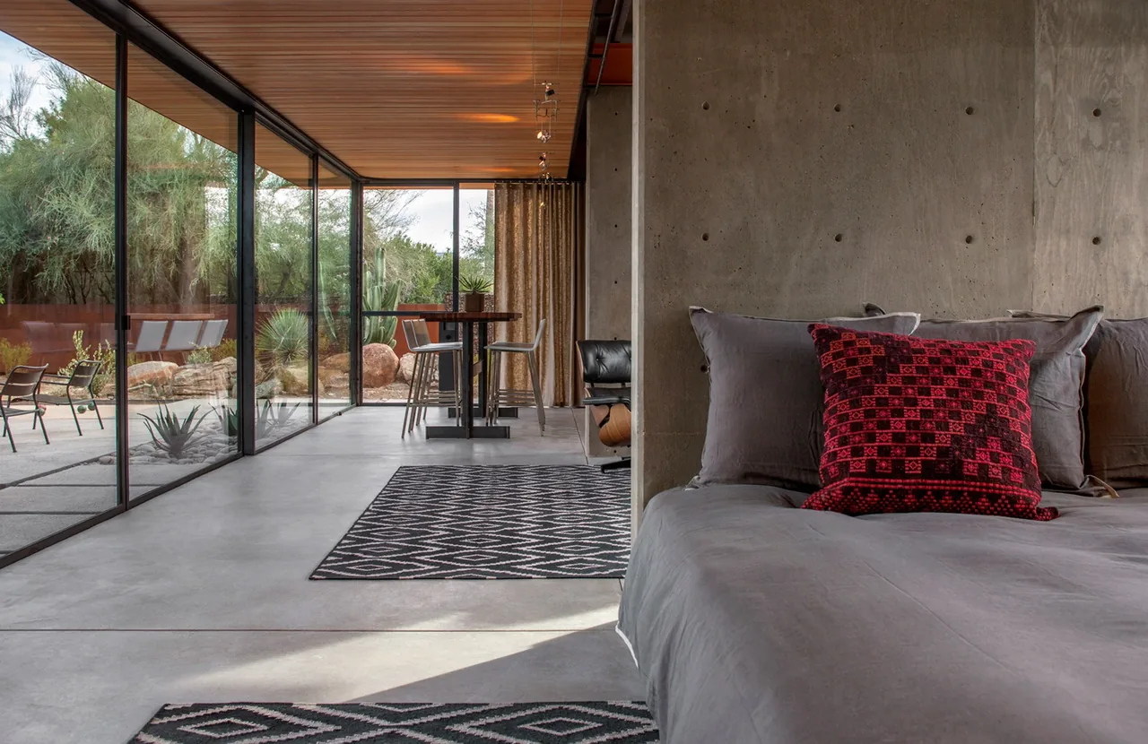 The barn guest house features a bedroom with a concrete wall and expansive floor-to-ceiling glass windows overlooking desert landscaping.
