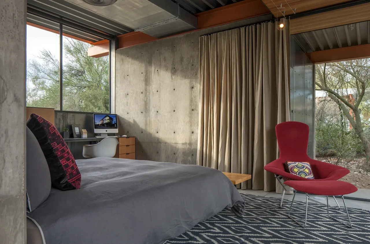 Bedroom in a barn guest house with concrete walls, large windows, and a red modern chair.