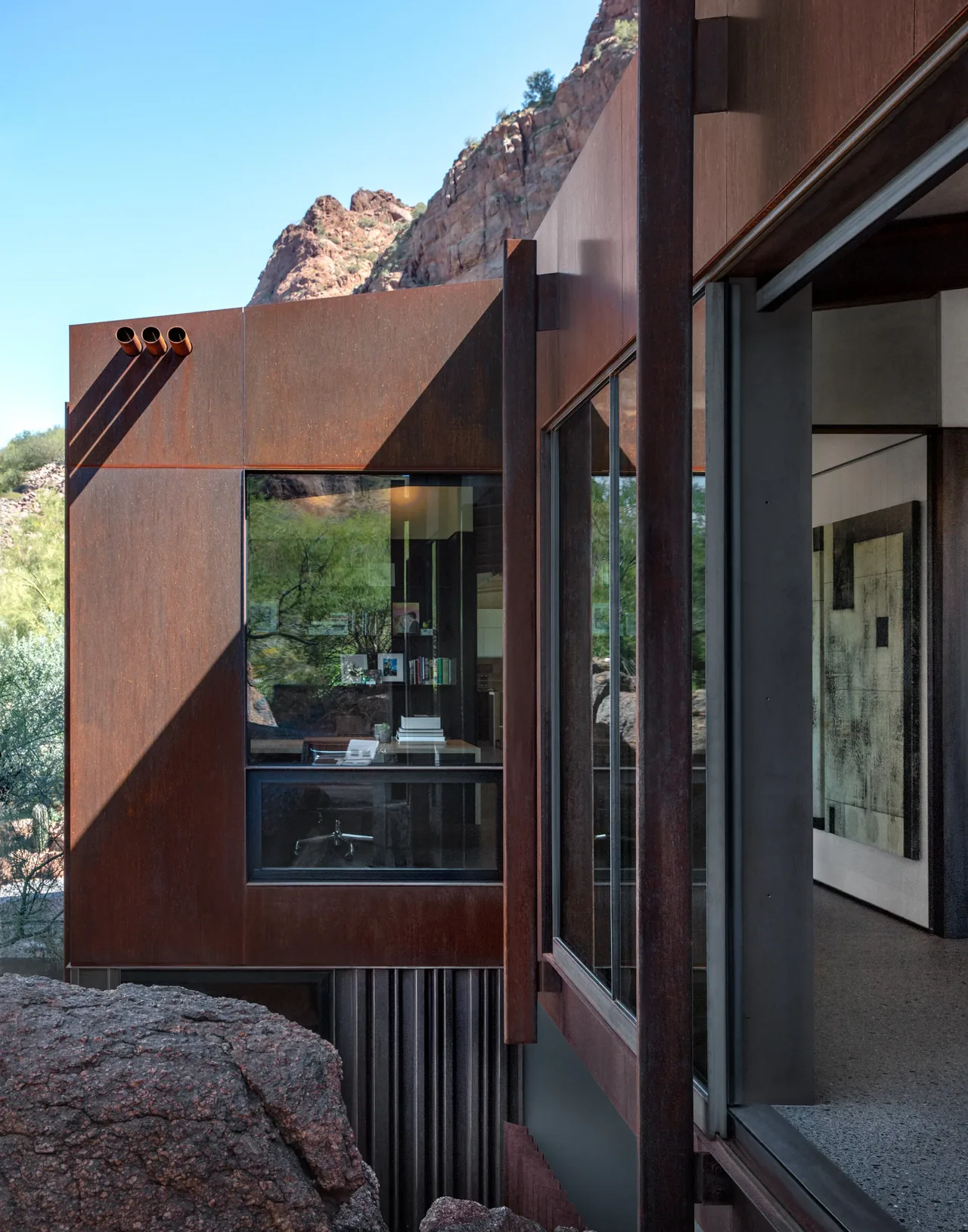 Modern dwelling with weathered steel facade and large glazed window overlooking desert rock formations in Phoenix.